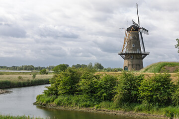 This scenic shot captures a classic Dutch windmill Gorinchem. standing tall against a cloudy sky, overlooking a tranquil waterway and lush green landscape. A symbol of the Netherlands.