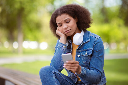 Thoughtful African American female student outdoors looking concerned at smartphone, upset black female seated on campus bench, reading message on mobile phone, got bad news