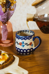 A beautifully patterned cup is filled with tea at a wooden table. Tea is being poured into a patterned teacup, a bouquet of flowers sits beside it. Tea party at home.