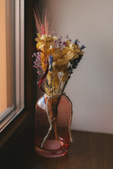 A beautiful display of dried flowers in a pink vase next to a window. A delicate dried flower arrangement in a rose-colored glass vase, set beside a window, creating a serene and cozy atmosphere.