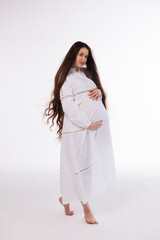 Pregnant woman in white long linen dress with very long hair on white background. Dreamy tender girl expecting a child. Portrait of beautiful pregnant woman in white shirt, smile.
