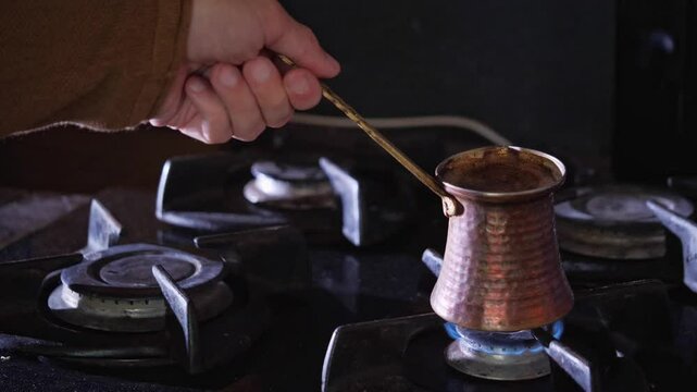 An authentic and ritualistic mood is captured as a hand holds a traditional copper cezve pot over a blue gas flame, actively brewing Turkish coffee. This shot is ideal for depicting cultural culinary 