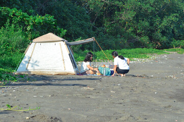 Two teenage girls, one squatting and one sitting, are organizing food and drinks on a mat under a canopy in front of a tent on a sandy beach near coastal plants and trees in the morning.