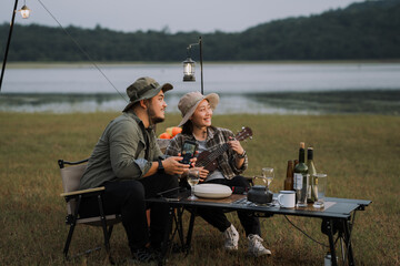 A man and a woman are sitting on a lawn near a lake. The man is holding a guitar and the woman is holding a wine glass. summer camping outdoor lifestyle concept.