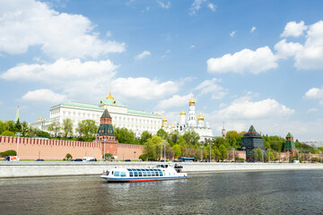 Fototapeta premium A scenic view of the Moscow Kremlin along the river. The historic architecture features towers and domes under a clear blue sky with fluffy clouds. A boat sails on the water.