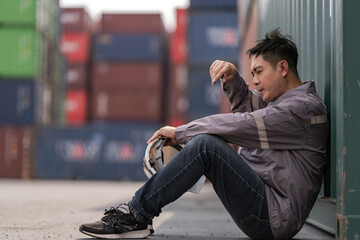 A tired logistics worker in a gray uniform sits on the ground at a busy shipping container yard,...