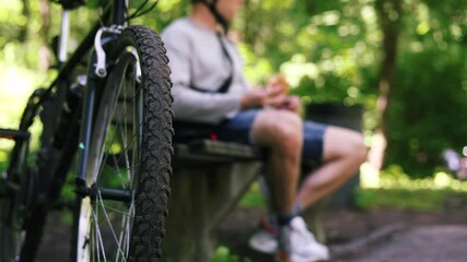 Young Caucasian man eating sandwich and drinking coffee while sitting on bench to resting after cycling in park. - Powered by Adobe