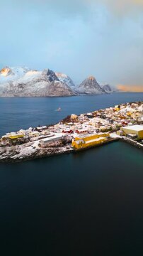 Fischerboot f&auml;hrt im Winter bei Sonnenaufgang an Hus&oslash;y bei Senja vorbei hinaus aufs offene Meer, schneebedeckte Gipfel im Hintergrund leuchten im warmen Licht &uuml;ber der Fjordlandschaft Nordnorwegens