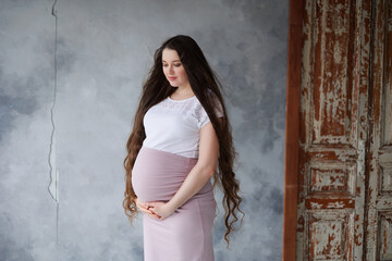 Fototapeta premium Pregnant woman in dress with very long hair on grey background. Dreamy tender girl expecting a child. Portrait of beautiful brunette pregnant woman. Window