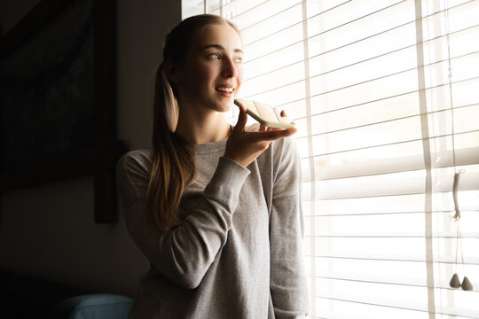 Portrait of young woman using voice assistant on smartphone at home smiling recording audio message near window blinds in living room making call talking on loudspeaker chatting in social network