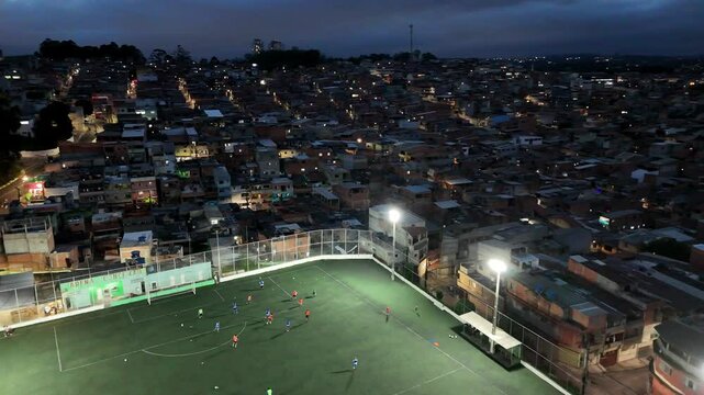 Drone view of a favela football pitch in Osasco, S&atilde;o Paulo, lit by floodlights with players on the field and houses packed tightly around it.