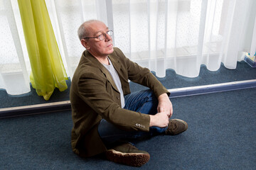 A middle-aged man in a green jacket sits on the floor near a panoramic window