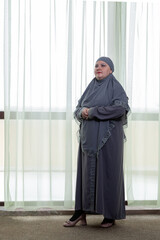 A Muslim woman in a modest gray hijab stands against a panoramic window