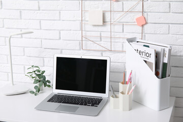Modern workplace with laptop, lamp, magazines and stationery near white brick wall