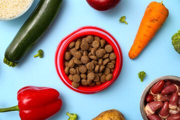 Bowl with healthy dry pet food near vegetables and meat on blue background