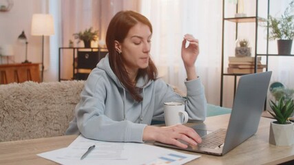 Exhausted lazy young woman freelancer working on laptop at table on couch looking tired at the end of workday holding head with one hand. Documents, cup of coffee are on table. Girl working from home. - Powered by Adobe