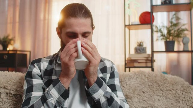 Young man holding steaming coffee or tea at home, closing eyes to soak smell in comforting fragrance. Guy on sofa inhales deeply, relishing each slow drinking sip mouthful with visible satisfaction.