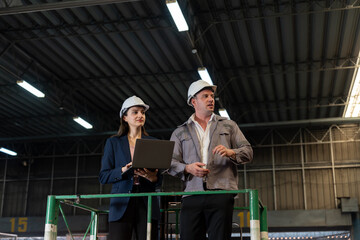 Naklejka premium Male and female engineers wearing white safety helmets standing on elevated platform inside warehouse. Woman holding laptop while discussing project with colleague in professional industrial 