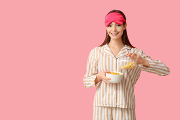 Young woman in pajamas pouring tasty cornflakes into bowl on pink background