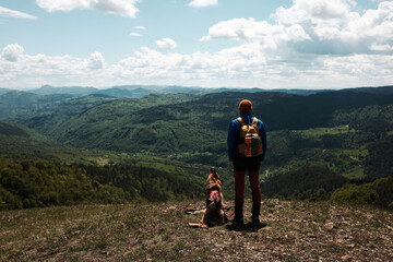 Male hiker and German Shepherd dog standing at a scenic viewpoint, gazing at the vast green valley...