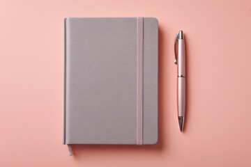 Neutral-toned writing set: a grey notebook with a matching elastic closure and a pink metallic pen sit on a pastel pink surface