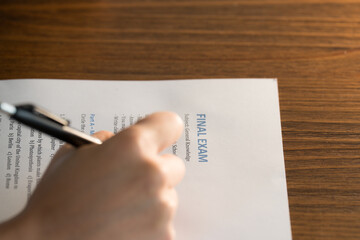 Student’s hand holding a black pen while taking a printed final exam on general knowledge. The paper rests on a wooden desk and the visible section shows the title “FINAL EXAM”