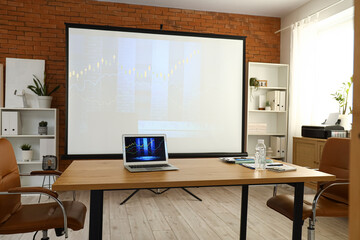 Laptop with diagrams of exchange rates on table and projector screen in office