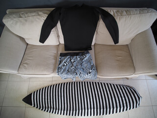 Black wetsuit and swim trunks lie on a couch next to surfboard, suggesting preparations for a surfing.