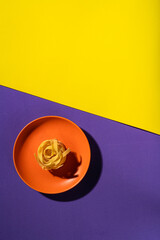 Single raw tagliatelle nest on orange plate over diagonal yellow and purple background. Minimalist...