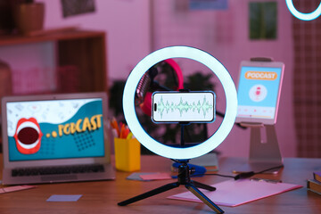 Mobile phone with voice recording and ring light on podcaster's table in dark studio, closeup