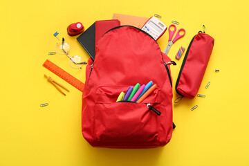 Red school backpack with different stationery supplies and eyeglasses on yellow background