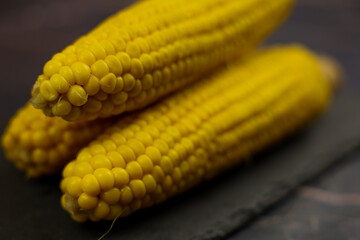 Three yellow peeled ears of corn with large grains lie on a black stone plank against a dark background in the center. The concept of healthy nutrition, natural iron, corn cultivation. High quality