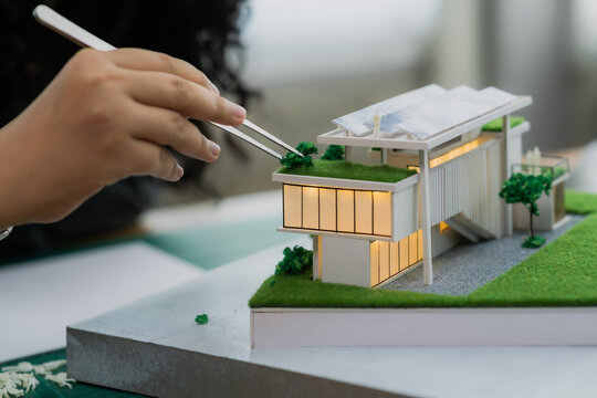 Using tweezers for precision, an architect adds miniature trees to the green roof of an illuminated, modern house model - Powered by Adobe