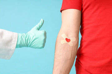 Male blood donor with medical patches and doctor's hand showing thumb-up on blue background, closeup