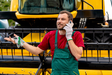 Smiling male farm worker talking on phone near combine harvester outdoors