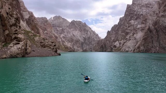 A lone kayaker paddles across the vibrant turquoise waters of Kel Suu Lake, surrounded by towering, rugged mountains under a cloudy sky