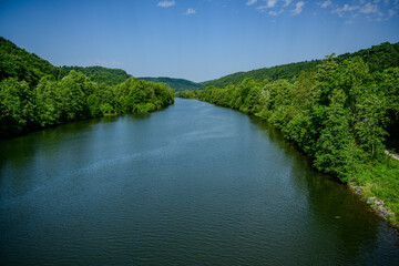 Fototapeta premium Serene river flows through lush green hills in a tranquil landscape with blue skies and fluffy clouds