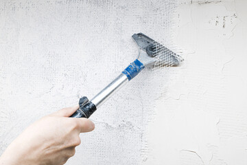 Close-up of a worker’s hand using a scraper tool to remove old plaster from an interior wall during home renovation. Shows rough surface texture and manual repair in progress