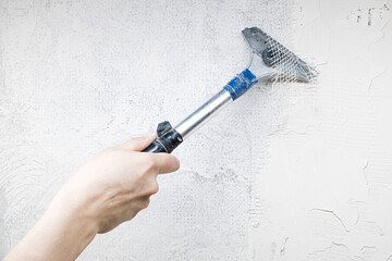 Close-up of a worker’s hand using a scraper tool to remove old plaster from an interior wall during home renovation. Shows rough surface texture and manual repair in progress