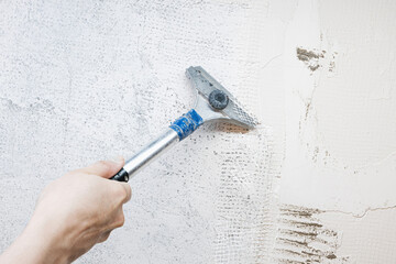 Close-up of a worker’s hand using a scraper tool to remove old plaster from an interior wall during home renovation. Shows rough surface texture and manual repair in progress
