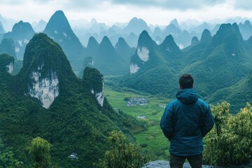 Obraz premium Back view of young male hiker in outdoor jacket standing on mountain top, overlooking green mountains in China, panoramic landscape, Asian travel adventure concept.