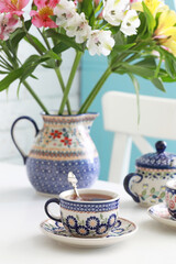 A beautiful ceramic cup and saucer with tea on a white table. A jug with flowers in the background. Selective focus.