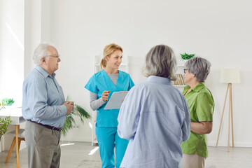 Fototapeta premium Group of elderly people listening young woman nurse or psychologist talking with seniors on a meeting standing in a circle in nursing home. Psychological support for retired people.