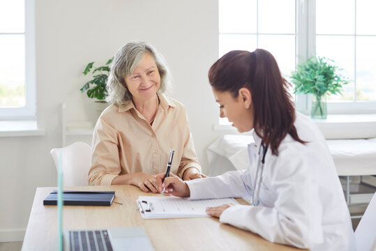 Young doctor writing out a prescription for a senior woman patient. Happy, smiling, old, gray haired lady sitting at a table with her health care provider and waiting for her medical prescription