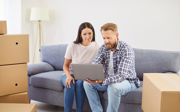 Happy young married couple man and woman sitting on sofa in living room in new apartments using laptop together with unpacking cardboard boxes. Moving day, relocation and rent concept.