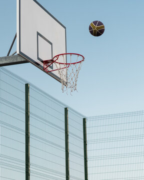 Photo of basketball ball in the air over blue sky background. The ball is about to hit the ring on the outdoor basketball court surrounded with netting.