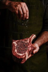 Close-up of a chef’s hands seasoning a raw tomahawk ribeye steak with coarse salt and pepper. The thick, marbled cut of meat is held in one hand, while the other sprinkles flaky sea salt in mid-air. 