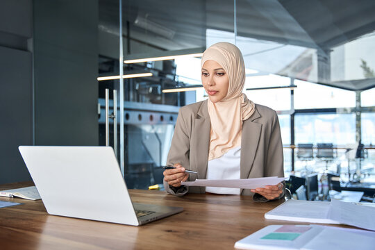 Executive Indian or arab businesswoman working with documents at desk in office. Middle eastern muslim woman in hijab for business analysing, trading, banking, financial data using laptop pc computer