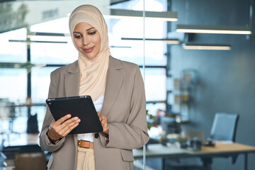 Focused young Indian or arab entrepreneur businesswoman holding digital pc tablet standing in modern office. Smiling middle eastern woman in suit and hijab working using touchpad computer. Copy space