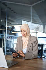 Middle Eastern muslim businesswoman manager ceo using cell phone mobile application. Smiling young indian woman in hijab holding smartphone sitting in office working online at desk on gadget. Vertical